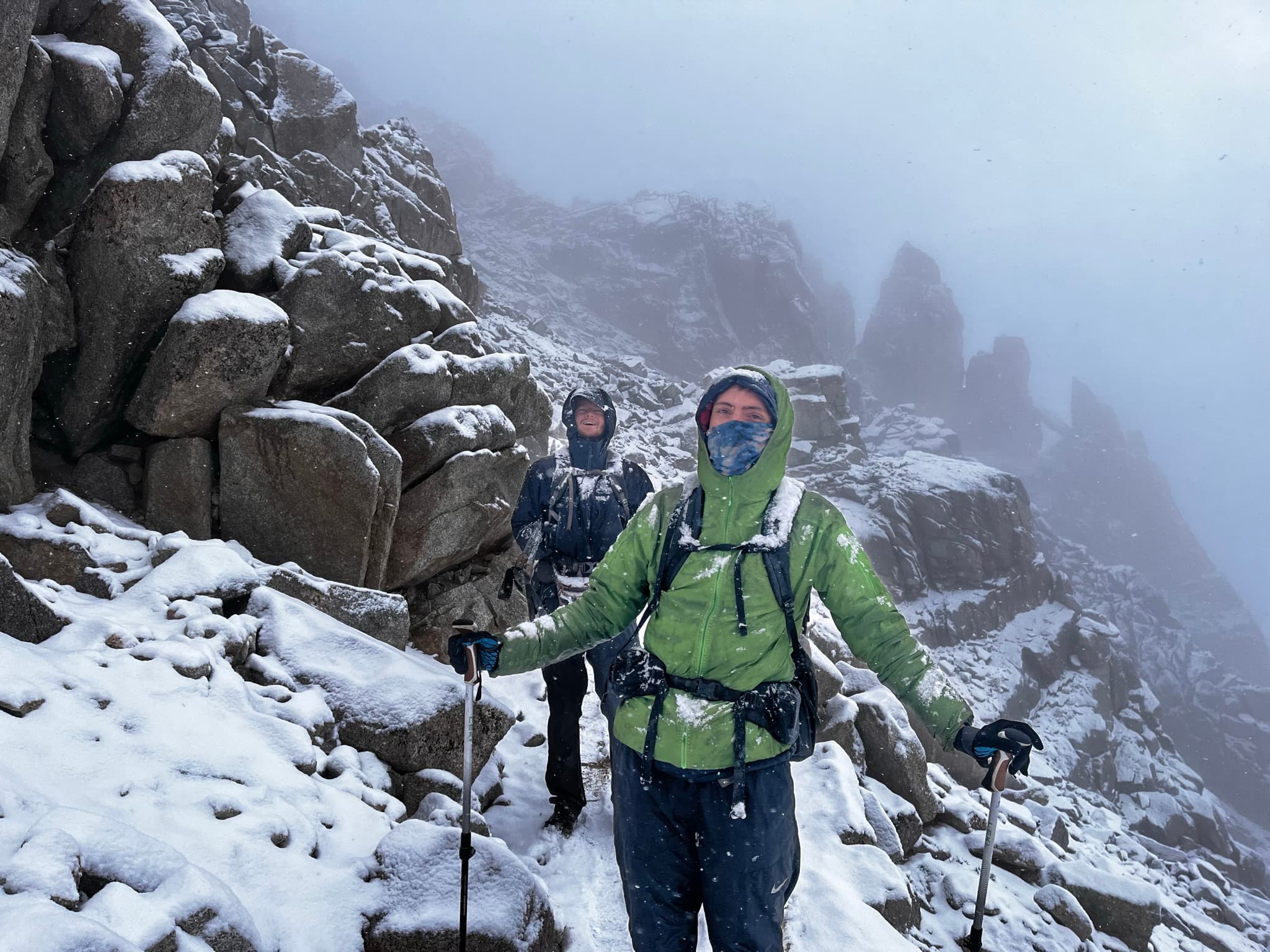 Two hikers bundled against snow on a rocky ridge with jagged spires behind