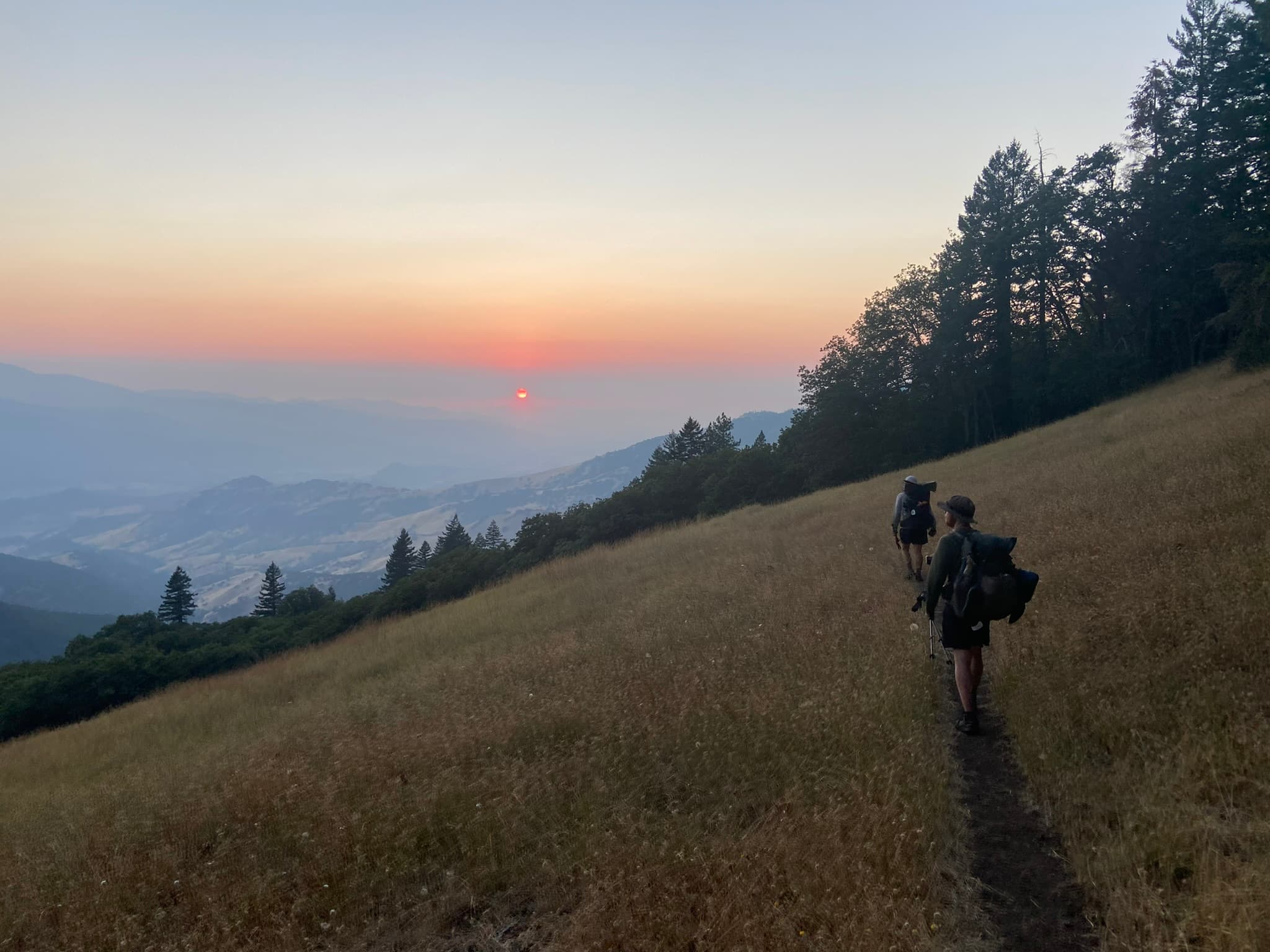 Hikers walking a golden grass ridge at sunset, smoke haze in the valley